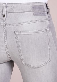 Jeans in denim grigio chiaro con design aderente. Caratteristiche include una tasca posteriore, cuciture sottili e una chiusura con bottone superiore. La texture appare liscia.