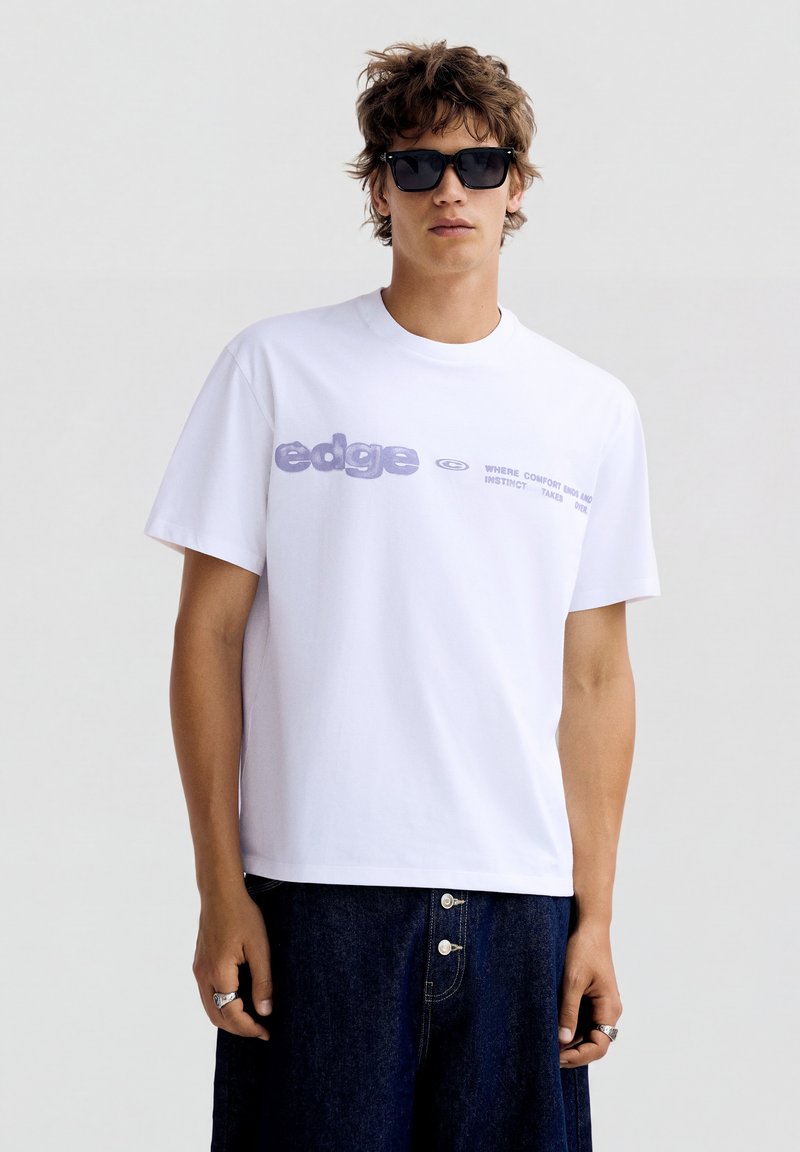 Young man with wavy hair wears black sunglasses, a white t-shirt with "edge" text and dark blue jeans with visible buttons.