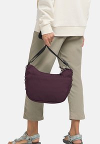 Lila Stoff-Schultertasche mit schwarzen Trägern, gewölbtem oberen Teil und gerafften Seiten. Person trägt einen hellbeigen Pullover und beigefarbene, Cropped-Hosen.