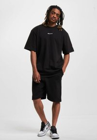 Juodas trumpas rankovėmis marškinius derinamas su juodais šortais. Marškiniai turi mažą logotipą ant krūtinės, o aprangą baigia balti ir juodi sportbačiai.