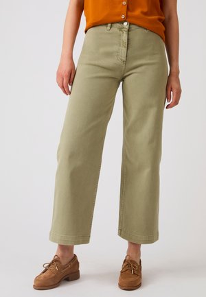 Persona che indossa pantaloni a gamba larga verde oliva, scarpe marroni stringate e una maglia arancione con bottoni, in piedi su uno sfondo uniforme.