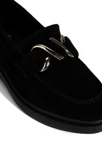 Black suede loafer with a textured surface, featuring a metallic decorative buckle, rounded toe, and a flat rubber sole.