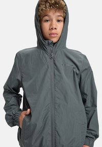 Lättviktsjacka med huva i mörkgrått tyg. Har elastiska manschetter, dragkedjestängning och sidofickor för förvaring. Slät, vattentålig yta.