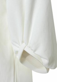 Weißes Baumwollhemd mit voluminösem Puffärmel, der ein geknotetes Detail am Bund und eine glatte, matte Textur aufweist.