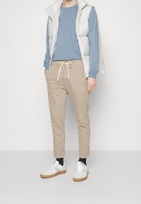 Lekka biała pikowana kamizelka na niebieskiej koszuli z długim rękawem, zestawiona z beżowymi spodniami ze sznurkiem i białymi trampkami na gumowej podeszwie.