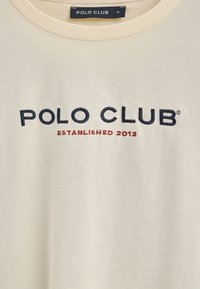 T-shirt color crema con scollatura a costine. Presenta la scritta ricamata "POLO CLUB" in blu navy e "ESTABLISHED 2012" in rosso. Tessuto morbido e leggero.