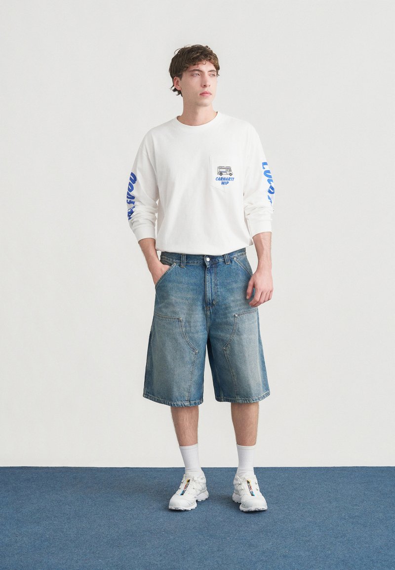 Hvid skjorte med lange ærmer, blå tekst og lomme; lyseblå denimshorts med to store forlommer; kombineret med hvide sneakers og sokker.