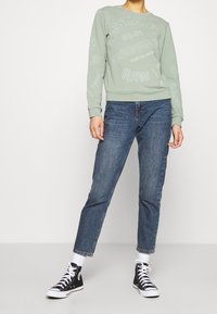 Sudadera verde con texto blanco, combinada con vaqueros de mezclilla azules y zapatillas altas negras. Diseño sencillo y casual con puños acanalados.