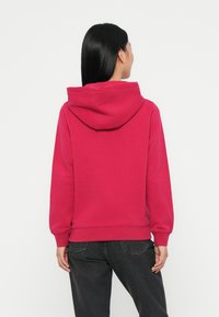 Sweatshirt à capuche fuchsia avec des bords côtes aux poignets et à l'ourlet. Texture lisse, coupe décontractée et une capuche spacieuse. Porté avec un pantalon gris foncé.