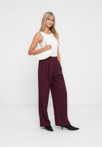 Weißes, enges Tanktop, burgunderfarbene weite Hose und eine weiße Handtasche mit Nieten-Tragegurt. Schwarze spitze Absätze runden das Outfit ab.