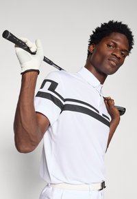 Pólo branco de manga curta com riscas pretas, usado por uma pessoa que segura um taco de golfe. O tecido é suave e apresenta um colarinho com botões.