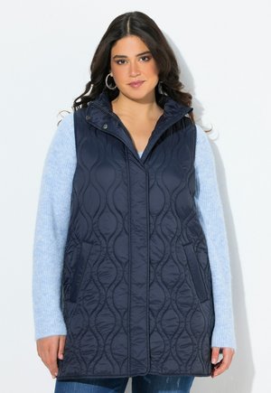 Gilet trapuntato blu navy con colletto alto, zip frontale e tasche laterali, sovrapposto a un maglione azzurro chiaro, caratterizzato da una trama liscia.