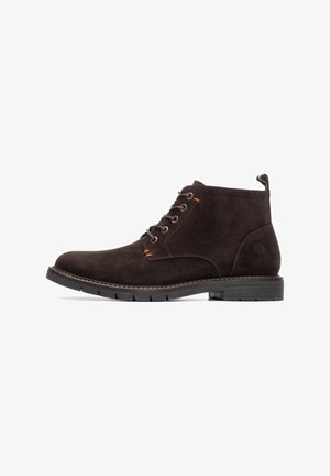 Botines de ante marrón con seis ojetes de metal, acentos en naranja contrastante, suela de goma gruesa y una cinta en la parte trasera.