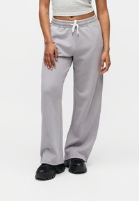 Άτομο που φοράει ανοιχτό γκρι φόρμα με κορδόνι και μαύρα chunky αθλητικά παπούτσια, đứng σε απλό λευκό φόντο.