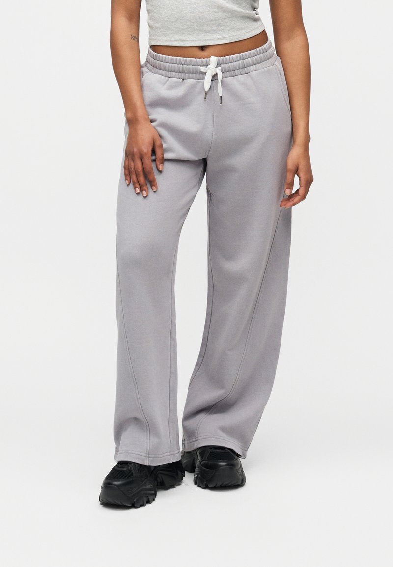 Άτομο που φοράει ανοιχτό γκρι φόρμα με κορδόνι και μαύρα chunky αθλητικά παπούτσια, đứng σε απλό λευκό φόντο.