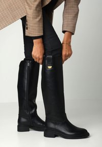 Kozačky z černé kůže po kolena s hladkou texturou, kulatou špičkou a nízkým blokovým podpatkem. Obsahují boční zip a zlaté kovové doplňky.