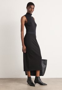 Robe sans manches noire avec un col haut, un corsage ajusté et une jupe évasée. Tissu texturé associé à des bottes noires à talons aiguilles et un sac fourre-tout.