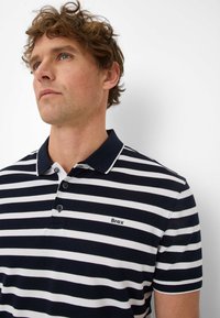 Navy and white striped polo shirt with a collar, featuring three buttons and a stitched brand logo on the left chest. Cotton material.