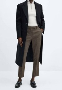 Manteau long noir sur un pull blanc côtelé, associé à un pantalon marron sur mesure et des mocassins noirs. Tissus texturés, lignes épurées.