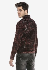 Schwarze und rote Jeansjacke mit einer distressierten Textur, silbernen Nieten auf den Schultern und einem taillierten Schnitt. Verfügt über eine Vorderreißverschlusstasche.