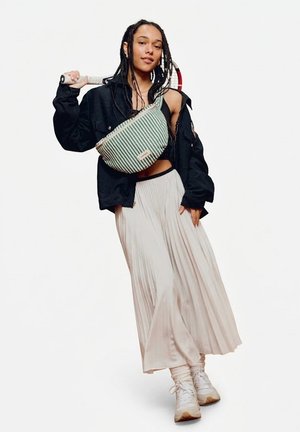 Femme aux cheveux tressés porte une veste noire, un sac bandoulière rayé, une jupe beige plissée et des baskets, tenant une raquette de tennis sur l'épaule.