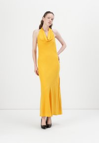 Robe jaune à encolure halter avec un décolleté drapé, coupe ajustée et ourlet évasé. Tissu lisse et extensible, associée à des talons hauts noirs.