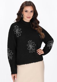 Zwarte trui met lange mouwen, hoge halslijn en decoratieve witte bloemenpatronen van kralen langs de voorkant en mouwen. Zachte textuur.