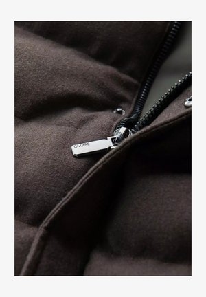 Veste rembourrée marron avec un tissu texturé, dotée d'une fermeture éclair noire et d'un tirette en métal argenté portant la mention "OMBRE". Gros plan sur le détail de la fermeture éclair.
