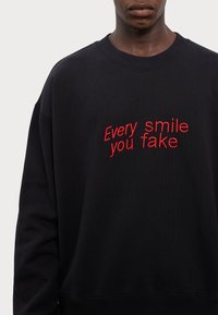 Sweat-shirt noir à col rond et manches longues, avec un texte brodé rouge : "Chaque sourire que tu fais" sur le devant.