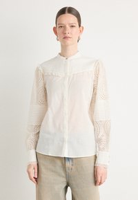 Camicia bianca con un busto texturizzato, maniche con un motivo simile al pizzo e chiusura con bottoni. Presenta un colletto alla cinese e un bordo arricciato.