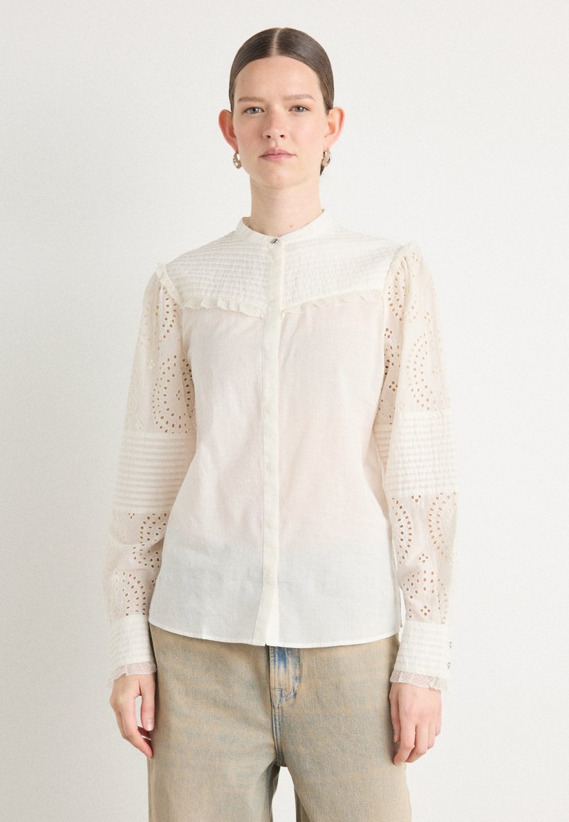 Camicia bianca con un busto texturizzato, maniche con un motivo simile al pizzo e chiusura con bottoni. Presenta un colletto alla cinese e un bordo arricciato.