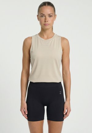 Vrouw die vooruit kijkt, gekleed in een beige mouwloos topje en zwarte strakke sportshorts tegen een witte achtergrond.