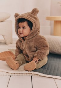 Sweat à capuche en polaire beige avec des oreilles d'ours, fermeture zippée et poignets côtelés. Pantalon vert olive et bottines marron doux avec des motifs clairs sur la semelle.