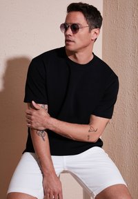 Juodas trumpomis rankovėmis marškinėliai su apvaliu iškirpimu, derinami su baltais šortais. Modelis dėvi apvalius akinius nuo saulės ir turi matomų tatuiruočių ant rankos.