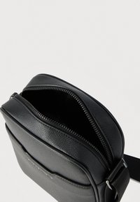 Μαύρο δερμάτινο τσαντάκι χιαστί με ανοικτό φερμουάρ και ρυθμιζόμενο λουρί σε απλό ανοιχτό φόντο.