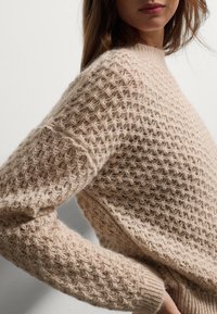 Maglione beige lavorato a maglia con un motivo geometrico testurizzato, maniche ampie e scollatura rotonda. Materiale morbido e peloso con polsini a coste.