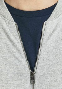 Närbild av en delvis zip-passad grå jacka över en mörkblå t-shirt med rund halsringning på en persons övre bröst.