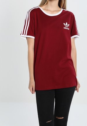 Vrouw draagt een bordeauxrode Adidas T-shirt met witte strepen op de mouwen en zwart gescheurde jeans, staand tegen een effen achtergrond.