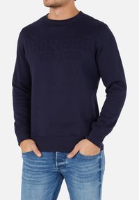 Mörkblå sweatshirt med rundad hals, texturerad logotyp "BOXEUR DES RUES", ribbade ärmslut och fåll, i kombination med ljusblå jeans.