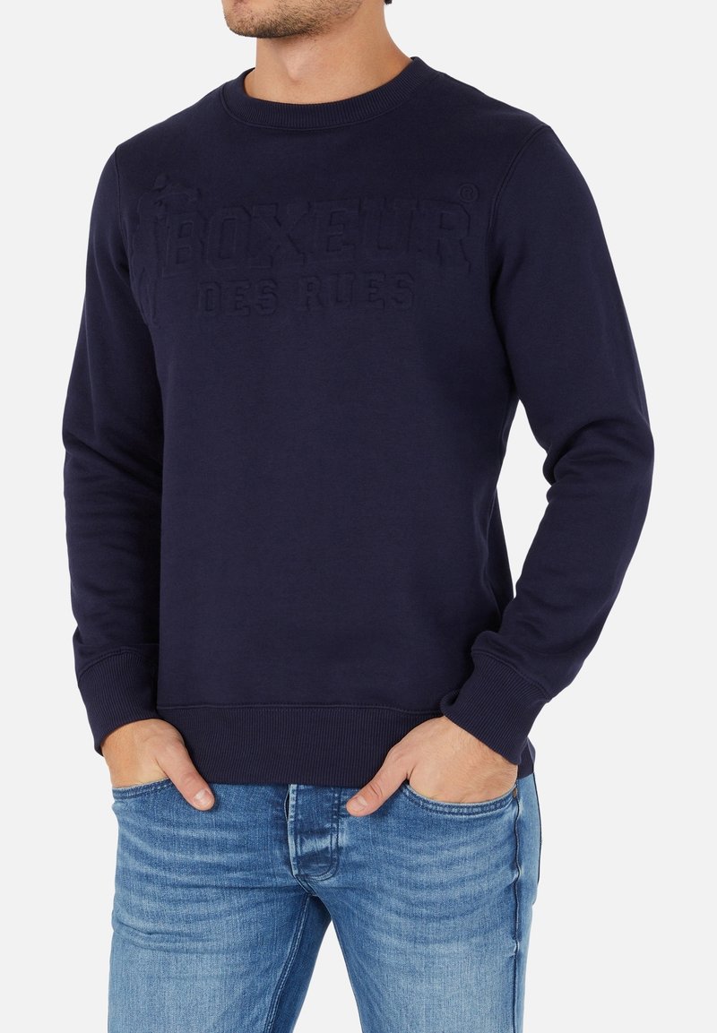 Mörkblå sweatshirt med rundad hals, texturerad logotyp "BOXEUR DES RUES", ribbade ärmslut och fåll, i kombination med ljusblå jeans.