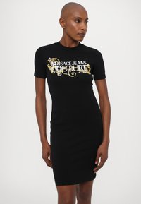 Juoda prigludusi suknelė iš medvilnės, su grafiniu spaudiniu, kuriame užrašyta "VERSACE JEANS COUTURE" baltomis ir auksinėmis spalvomis. Trumpomis rankovėmis, iki kelių ilgio dizainas.