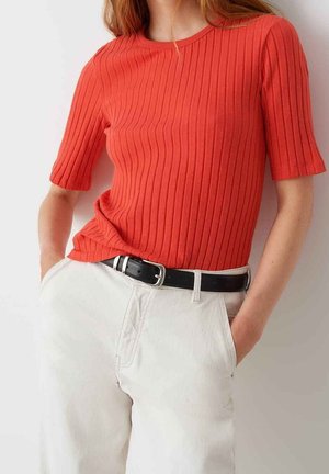 Gestreepte oranje top met korte mouwen, ronde halslijn, gedragen over witte broeken, aangevuld met een zwarte riem. Eenvoudig, getextureerd ontwerp.