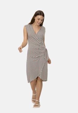 Mujer caminando con vestido cruzado sin mangas hasta la rodilla con patrón de zigzag multicolor y sandalias planas beige.