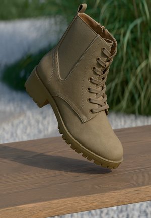 Beige suède veterschoen tot de enkel met dikke zool, in balans op een houten oppervlak, met vage groene planten en een achtergrond van witte grind.