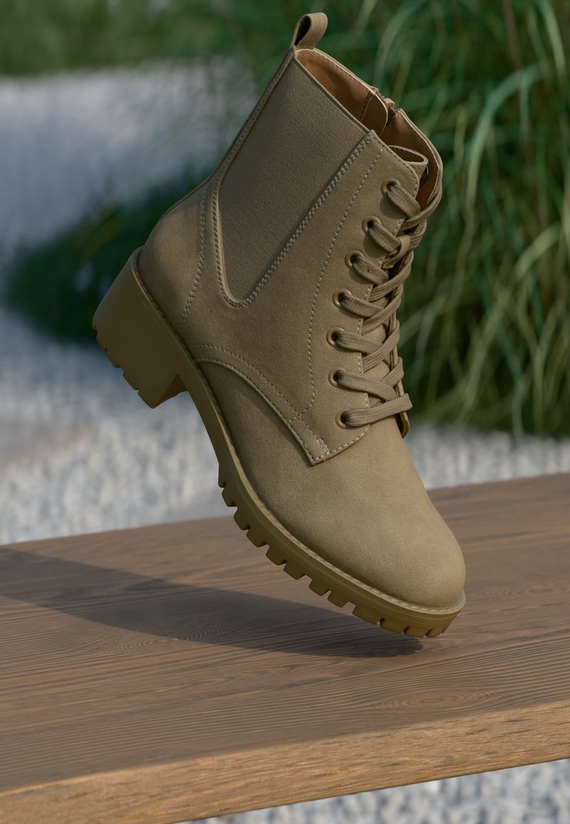Bota de tornozelo em camurça bege, com atacadores e sola chunky, equilibrada sobre uma superfície de madeira, com um fundo desfocado de plantas verdes e pedras brancas.