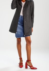 Abrigo de lana gris con solapas, cierre de botones, combinado con una falda vaquera azul y tacones de charol rojo con correas en el tobillo.