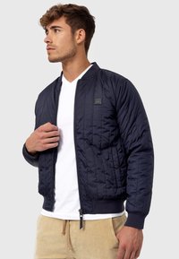 Marineblå quilted bomberjakke med ribbede manchetter og kant, lynlås lukning og et logo patch på brystet. Båret over en hvid V-hals skjorte.