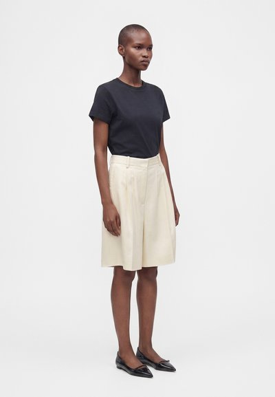 Femme aux cheveux courts, debout, portant un haut noir à manches courtes, une jupe plissée crème mi-longue aux genoux et des ballerines noires pointues sur un fond uni.
