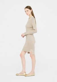 Abito beige a coste con una vestibilità aderente, maniche lunghe e colletto bianco. Presenta uno spacco laterale ed è abbinato a ballerine dorate.