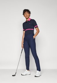 Námořnický golfový outfit s krátkými rukávy, který má růžový pruh přes hruď. Spárováno s námořnickými kalhotami a bílými sportovními botami. Golfová hůl v ruce.